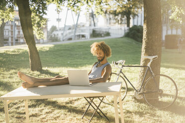 Man with beard and curly hair using laptop at table in park - KNSF01748