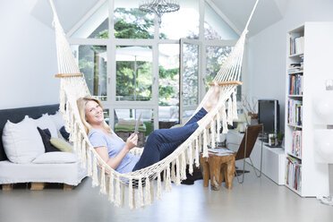 Lächelnde blonde Frau mit Tablet entspannt in der Hängematte im Wohnzimmer - MAEF12257