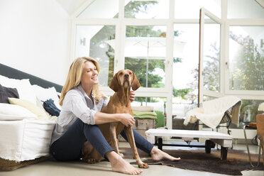 Relaxed woman sitting with her dog on floor of the living room - MAEF12263