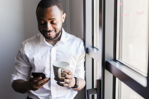 Smiling man holding cell phone and cup of coffee at the window stock photo