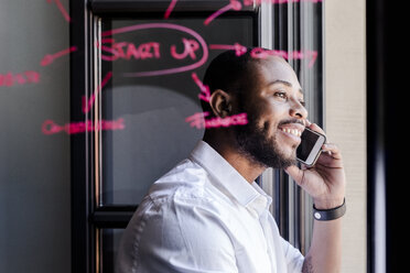 Smiling businessman on cell phone in office with writing on windowpane - GIOF02937