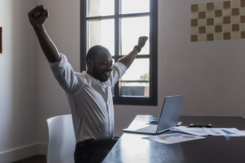 Happy businessman with laptop at desk raising his arms stock photo