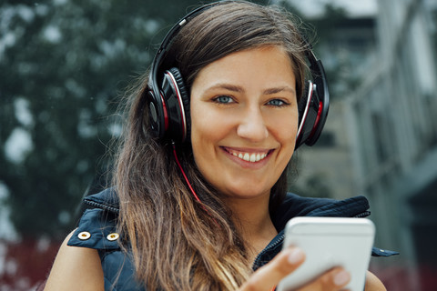 Porträt einer lächelnden jungen Frau mit Kopfhörern und Mobiltelefon in der Stadt, lizenzfreies Stockfoto