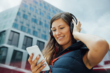 Lächelnde junge Frau mit Kopfhörern und Mobiltelefon in der Stadt - CHAF01913