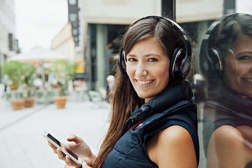 Porträt einer lächelnden jungen Frau mit Kopfhörern und Mobiltelefon in der Stadt - CHAF01915