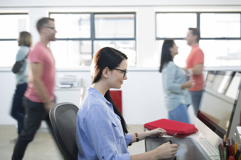 Employee working on her computer while other staff members walking past - ZEF14114