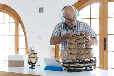 Älterer Mann mit Kopfhörern arbeitet an einem Schiffsmodell auf einem Tisch mit Tablet - JRFF01406