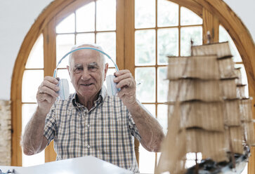 Porträt eines selbstbewussten älteren Mannes, der Kopfhörer aus einem Paket mit einem Schiffsmodell auf dem Tisch auspackt - JRFF01411
