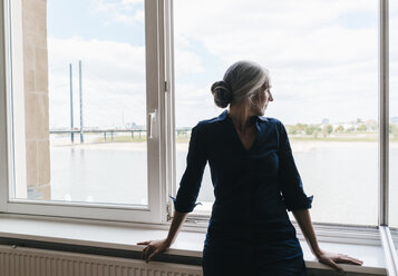 Businesswoman looking out of window in waterfront office - KNSF01762