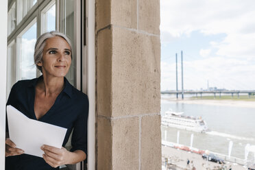 Lächelnde Geschäftsfrau schaut aus dem Fenster in einem Büro am Wasser - KNSF01771