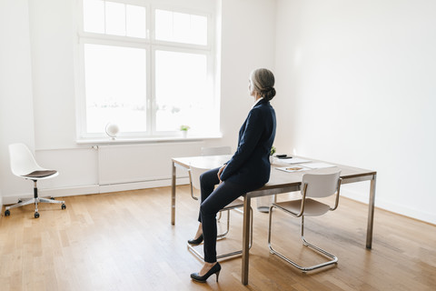 Geschäftsfrau sitzt auf einem Tisch im Büro und schaut aus dem Fenster, lizenzfreies Stockfoto