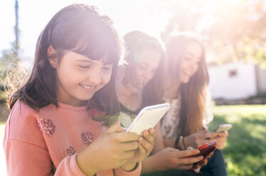 Drei glückliche Mädchen benutzen ihre Smartphones im Freien - MGOF03436