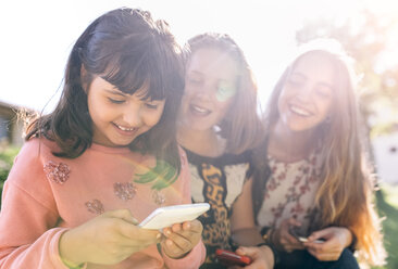 Drei glückliche Mädchen benutzen ihre Smartphones im Freien - MGOF03437