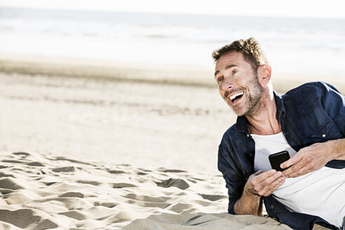 Glücklicher Mann mit Mobiltelefon am Strand - FMKF04205