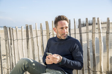 Man with cell phone sitting at fence on the beach - FMKF04210