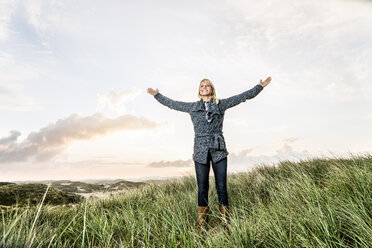 Happy woman standing in dunes with raised arms - FMKF04220