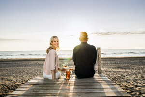 Paar mit Weingläsern auf der Promenade am Strand bei Sonnenuntergang sitzend - FMKF04242