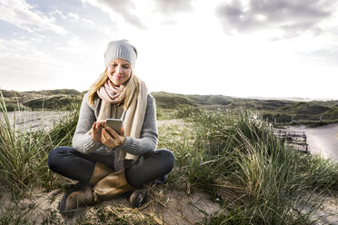 Smiling woman sitting in dunes using cell phones - FMKF04259