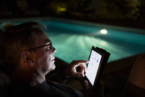 Man using tablet at the poolside at night - MAEF12357