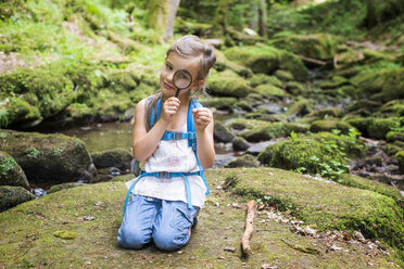 Kleines Mädchen mit Lupe, das auf einem Felsen im Wald hockt und eine Feder beobachtet - DIKF00266
