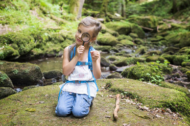 Little girl with magnifier crouching on rock in the woods watching a feather - DIKF00266