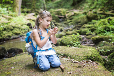 Kleines Mädchen mit Lupe, das auf einem Felsen im Wald hockt und eine Feder beobachtet - DIKF00267