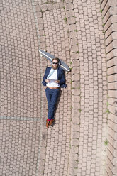 Businessman with skateboard lying on a wall using tablet and headphones - MAEF12358