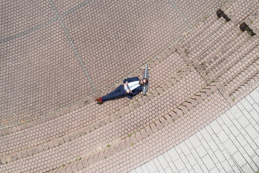 Businessman with skateboard lying on a wall using tablet and headphones - MAEF12359