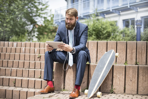 Geschäftsmann mit Skateboard, der ein Tablet und Kopfhörer benutzt - MAEF12363