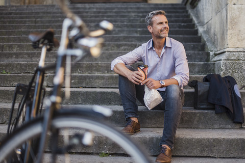 Lächelnder Mann auf einer Treppe sitzend mit Kaffee zum Mitnehmen in der Stadt, lizenzfreies Stockfoto