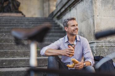 Lächelnder Mann auf einer Treppe sitzend mit Croissant und Kaffee zum Mitnehmen in der Stadt - DIGF02566