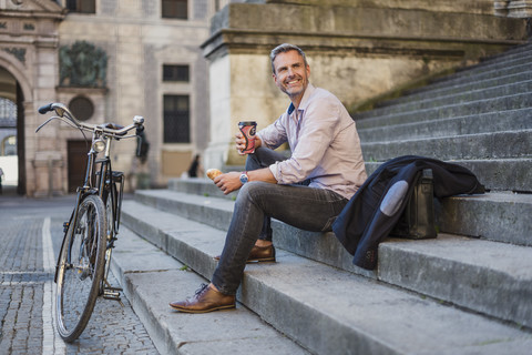 Lächelnder Mann auf einer Treppe sitzend mit Kaffee zum Mitnehmen in der Stadt, lizenzfreies Stockfoto