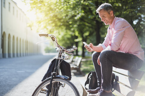 Smiling man with bicycle checking the phone on a park bench - DIGF02570