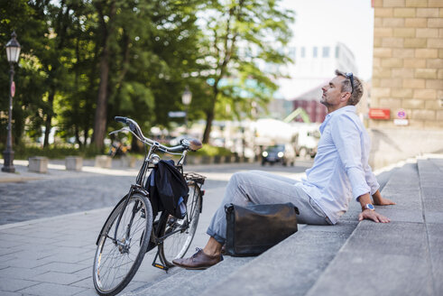 Entspannter Mann sitzt auf einer Treppe in der Stadt neben einem Fahrrad - DIGF02587