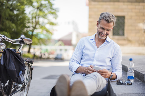 Lächelnder Mann sitzt auf einer Treppe in der Stadt und benutzt ein Mobiltelefon, lizenzfreies Stockfoto