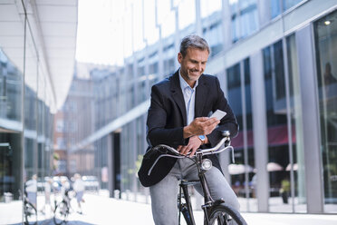 Smiling businessman with bicycle and cell phone in the city - DIGF02598