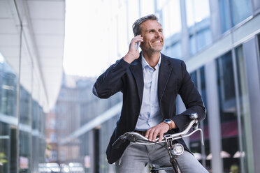 Smiling businessman with bicycle on cell phone in the city - DIGF02599