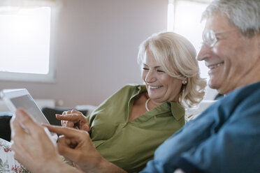 Happy senior couple at home sitting on couch using tablet - ZEDF00778