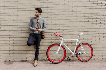 Porträt eines jungen Mannes mit Rennrad und Handy an die Wand gelehnt - GIOF02966