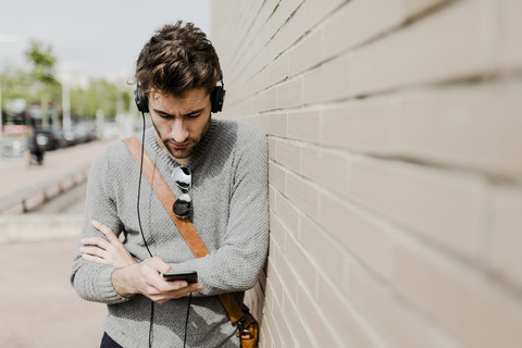 Junger Mann mit Kopfhörern lehnt an der Wand und schaut auf sein Handy, lizenzfreies Stockfoto