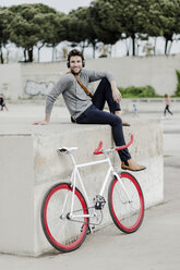 Young man with racing cycle sitting on a wall listening music with headphones - GIOF02976