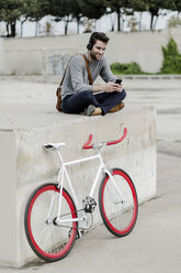 Young man with racing cycle sitting on a wall listening music with headphones - GIOF02977