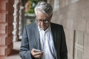 Älterer Geschäftsmann mit Kopfhörern, der auf sein Mobiltelefon schaut - GUSF00053