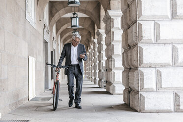 Älterer Geschäftsmann schaut auf sein Handy, während er sein Fahrrad schiebt - GUSF00056