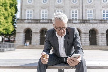 Älterer Geschäftsmann mit Eistüte, der auf einer Bank sitzt und auf sein Smartphone schaut - GUSF00062