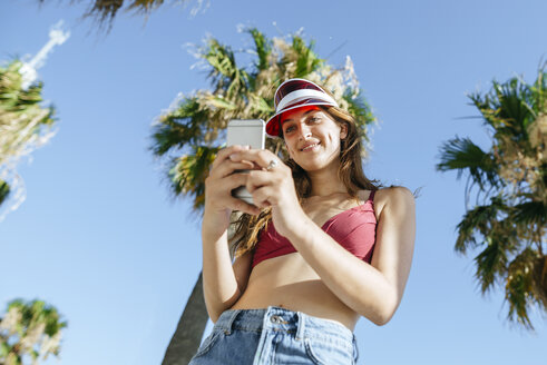 Junge Frau mit Sonnenblende und Bikini, die ein Mobiltelefon benutzt - KIJF01655