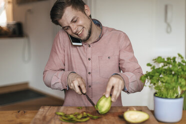 Smiling man on the phone chopping acocado in the kitchen - GUSF00063