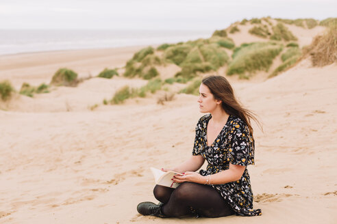 Nachdenkliche junge Frau sitzt mit einem Buch am Strand - NMSF00130