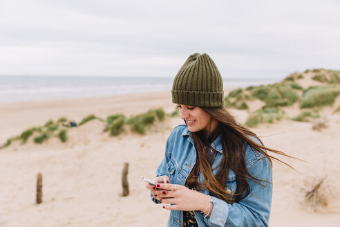 Lächelnde junge Frau, die am Strand auf ihr Handy schaut - NMSF00131