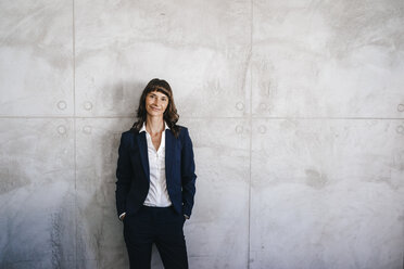Businesswoman leaning against office wall with hands in pockets - KNSF02006